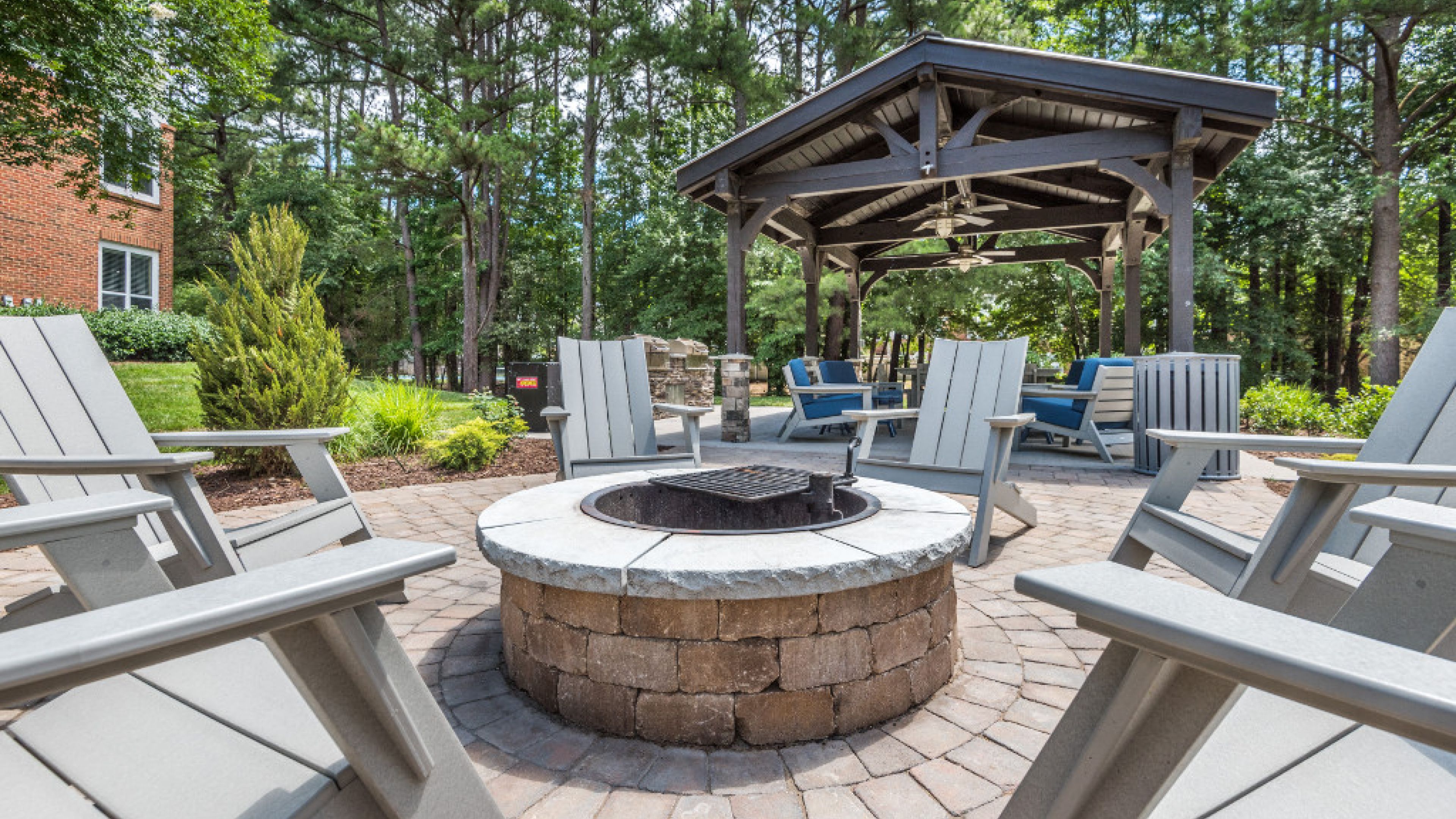 Hawthorne at the Parkway outdoor fire pit area with surrounding Adirondack chairs under a wooden gazebo, nestled among tall trees.
